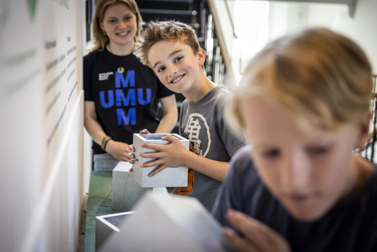 Twee jongens in museum houden kubus vast en luisteren er naar. Museumbegeleider (kennismaker) staat er naar te kijken. 